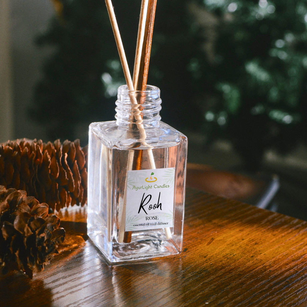 Clear diffuser bottle with reeds on a wooden surface, surrounded by pine cones and a blurred plant in the background.