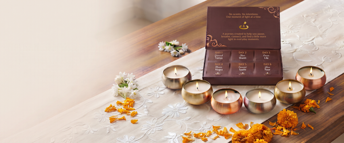 A set of six marigold tea light candles arranged around an open dark brown gift box on a white embroidered runner, with orange flower petals scattered on a wooden surface.